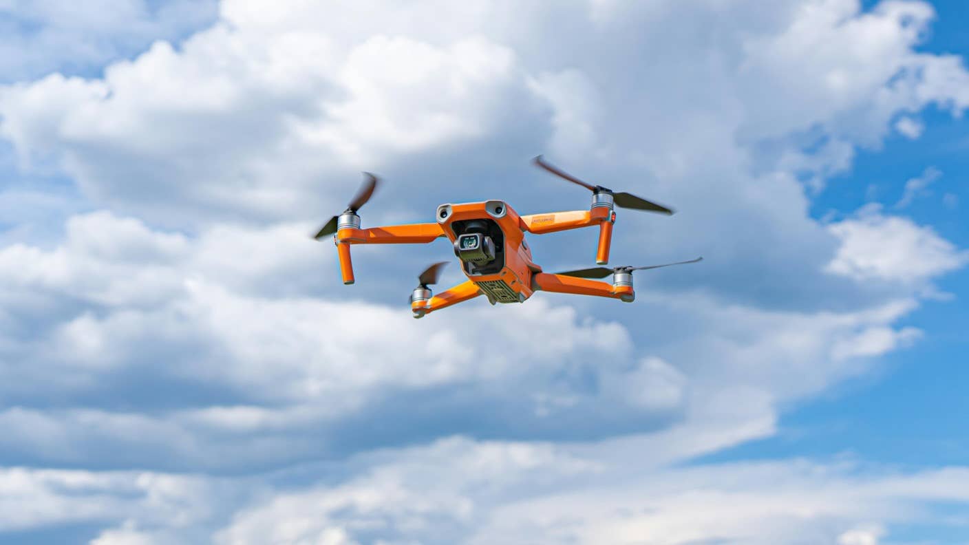 An orange drone is hovering in a blue sky with white clouds.
