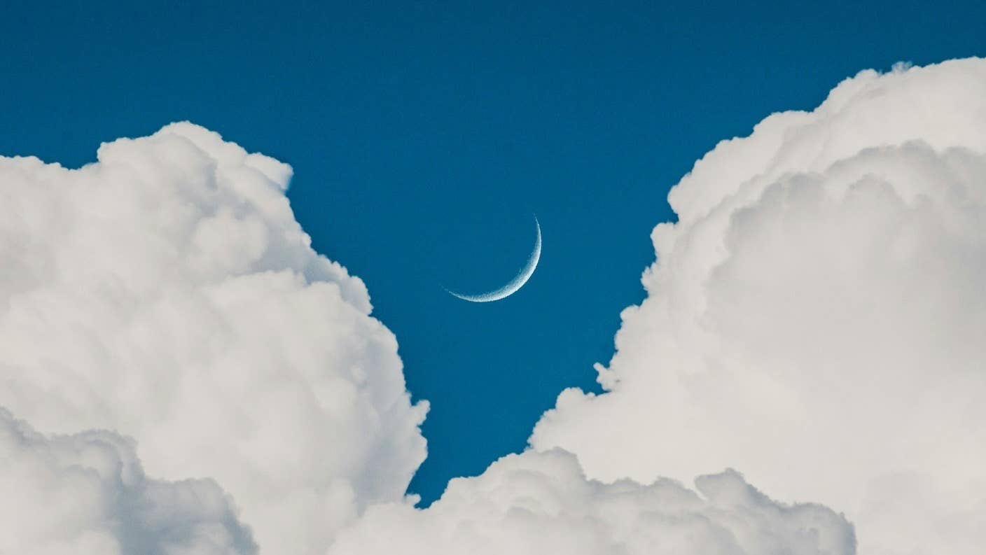 A fingernail moon peeks out between white clouds in a blue evening sky