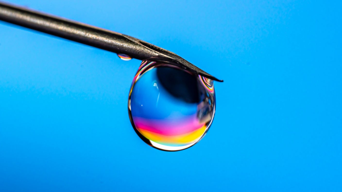A colorful drop of liquid is balanced on the tip of a needle over a blue background